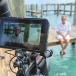 A camera monitor showing a man wearing sunglasses and a white shirt as he sits on a jetty. The same man is seen in the blurred background