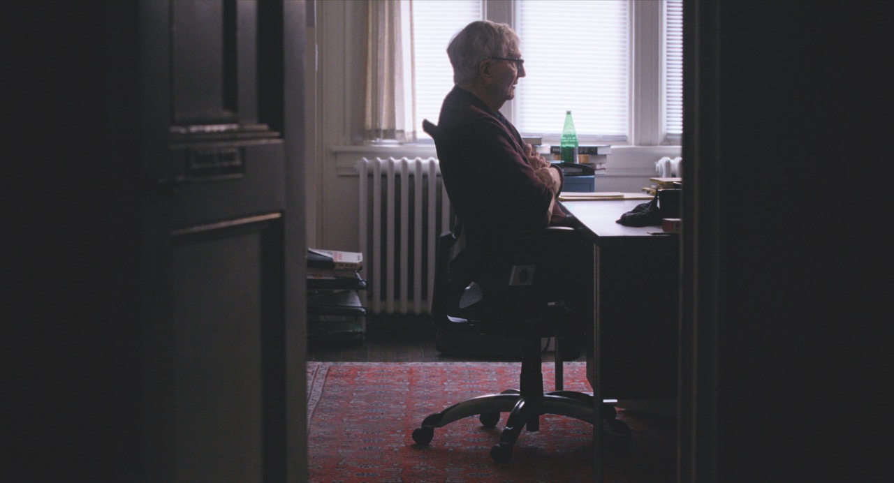 An older man sitting at a desk through an open door in a dimly lit office