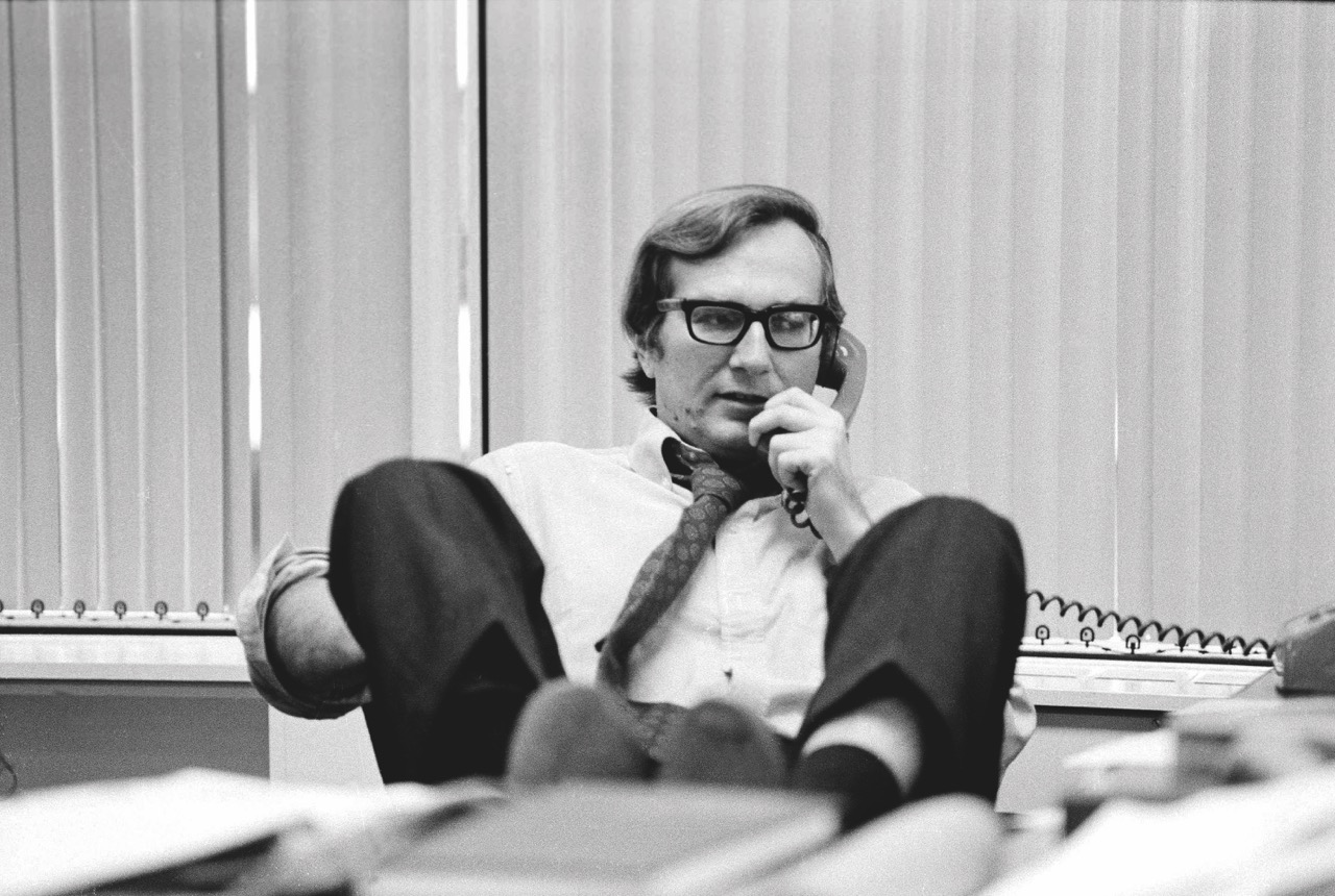 A black and white still of a person in an office using a telephone with their legs resting on the edge of a desk
