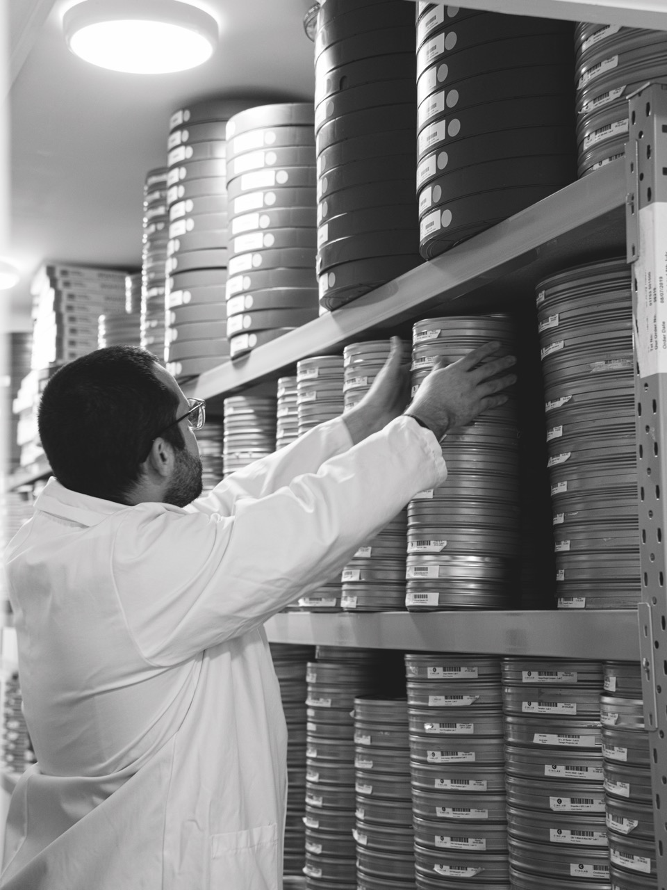 A black and white still of a man in a lab coat adjusting a stack of film on a shelf