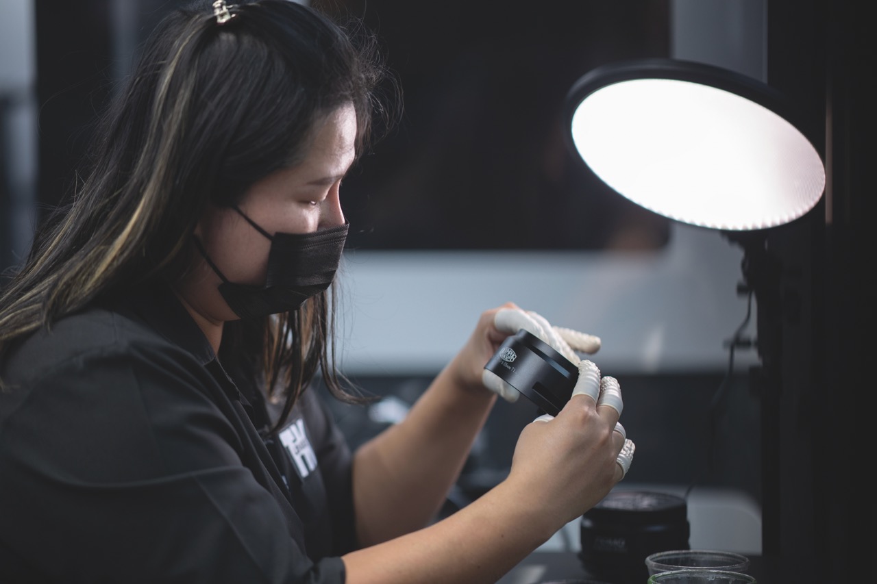 A person in a black mask and white tape on their fingers adjusting a lens under a light