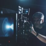 A man looking through a camera with a large lens attached, placed in a dark room