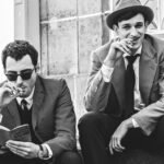 A black and white still of two men sitting on steps, wearing suits and smoking cigarettes. Another man in the background walks away