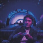 A young man wearing headphones and holding a glowing remote in a dark room