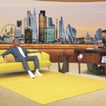 Two people on a talk show with a London skyline behind a yellow sofa