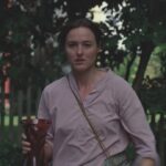 A person with a bag slung across their body, holding a glass, walking through a forested area