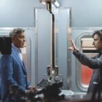 Two actors in suits standing opposite each other in a toilet, surrounded by cameras. One man has their arms raised